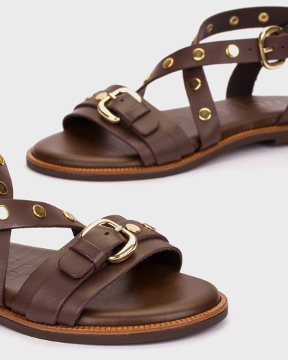 Detalle de las sandalias KORA 42 en piel marrón con remaches dorados hebilla decorativa y tiras cruzadas.